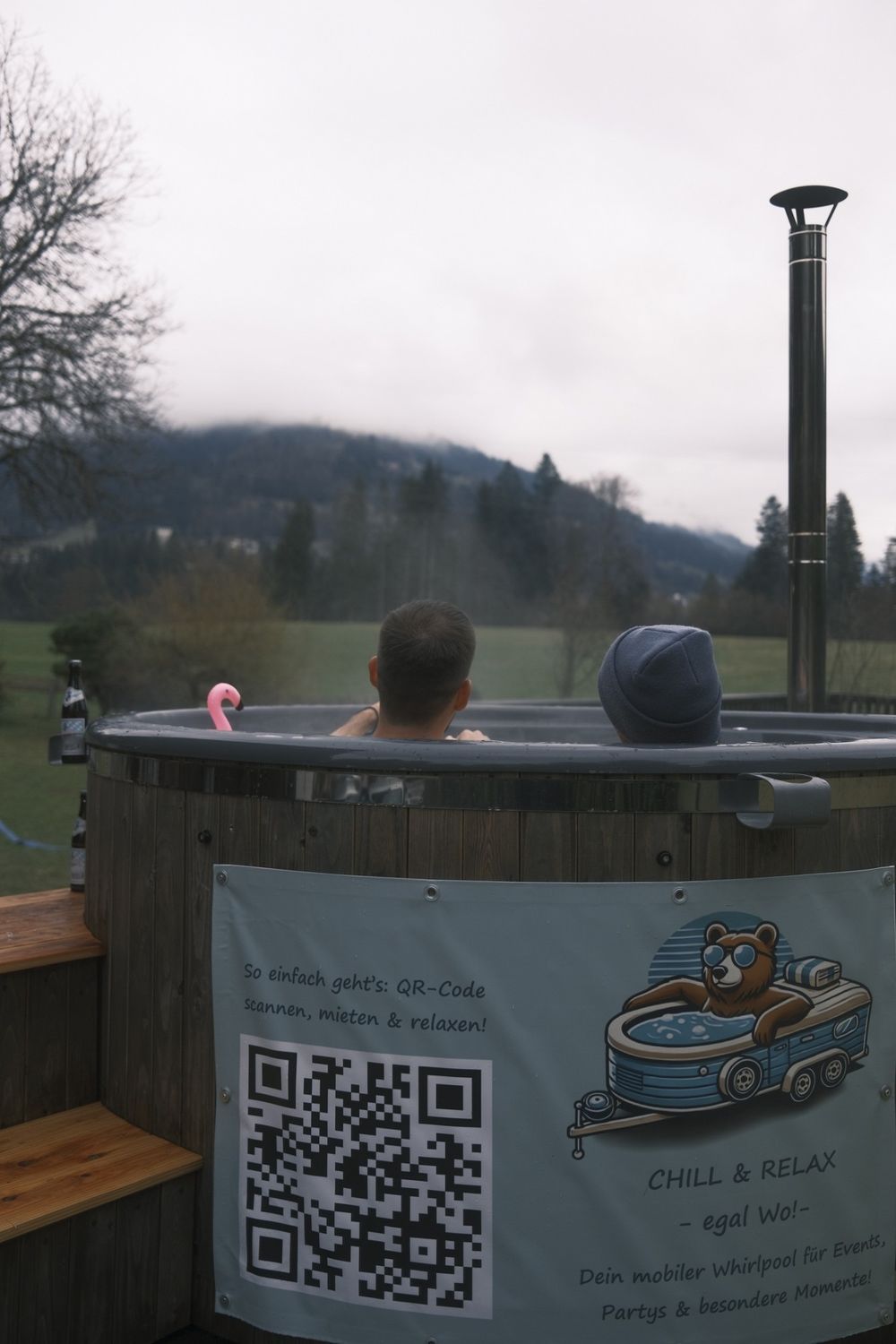 Zwei Personen entspannen im Holz-Whirlpool mit Blick auf Wiesen und Berge